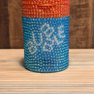 May include: A cylindrical container covered in small, sparkling rhinestones. The top section features orange rhinestones, while the bottom section is blue with the word BUBBLE spelled out in white rhinestones across the center of the blue surface.