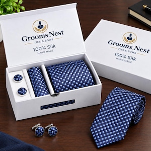 Può includere: Un set composto da cravatta, pochette e due paia di gemelli in seta con motivo a quadri blu e azzurro, presentati in una scatola bianca marchiata Grooms Nest Ties & Bows. Il set presenta un motivo intrecciato coerente, con il logo e la scritta 100% Silk Hand Made sul coperchio.