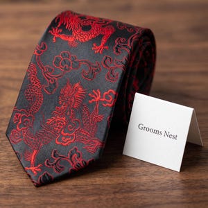May include: A black necktie featuring a vibrant red embroidered dragon pattern and floral motifs. The tie is folded and positioned next to a small white folded card with the text Grooms Nest printed in black on the front surface of the card.