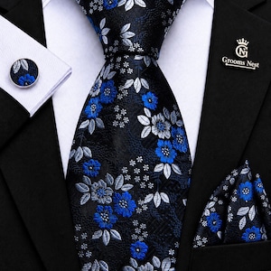 Peut inclure: Un ensemble composé d'une cravate, d'une pochette et d'un bouton de manchette noirs à motif floral bleu vif et gris argenté. La cravate est nouée sur une chemise blanche et un blazer noir. Une épingle argentée avec le texte Grooms Nest est fixée.