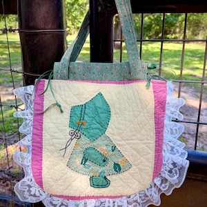 May include: A handmade quilted tote bag with a vintage aesthetic. The bag features a central appliqué of a bonneted figure in teal and floral fabrics, framed by pink and cream borders, and edged with white lace. The handles are made of a teal floral fabric.