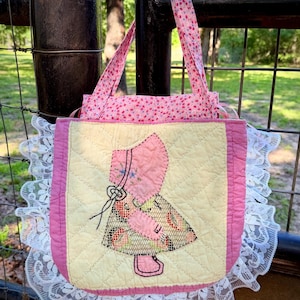 Puede incluir: Un bolso tote acolchado rosa y amarillo hecho a mano con un diseño de gorro. El bolso presenta un ribete de encaje blanco y asas con estampado floral rosa. El bolso está diseñado con un patrón acolchado y es perfecto para llevar objetos pequeños.