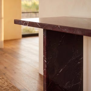 May include: A console table constructed from deep burgundy marble with prominent white veining. The piece features a thick, rectangular top surface supported by a vertical slab base, showcasing the natural polished stone texture and pattern.