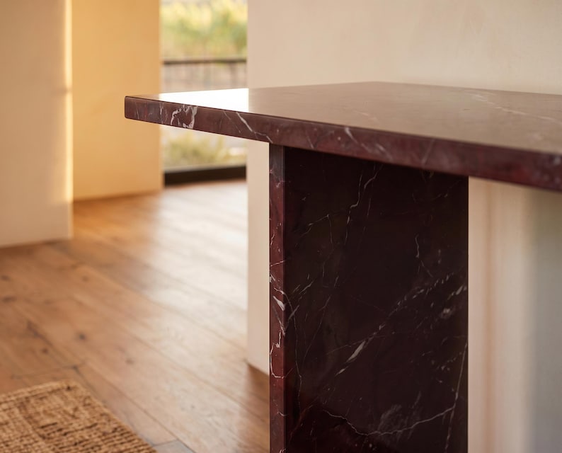 May include: A console table constructed from deep burgundy marble with prominent white veining. The piece features a thick, rectangular top surface supported by a vertical slab base, showcasing the natural polished stone texture and pattern.