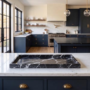 May include: A rectangular black marble tray with prominent white veining, featuring a smooth, polished surface and raised edges, positioned on a white marble countertop in a kitchen with dark blue cabinetry and brass hardware accents.