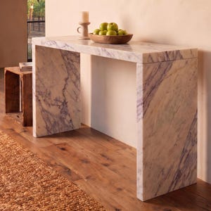 May include: A rectangular console table constructed from white marble with prominent grey and purple veining. The table features a waterfall edge design with two vertical side panels supporting a flat top surface, creating a clean, geometric silhouette.