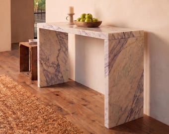 May include: A rectangular console table constructed from white marble with prominent grey and purple veining. The table features a waterfall edge design with two vertical side panels supporting a flat top surface, creating a clean, geometric silhouette.