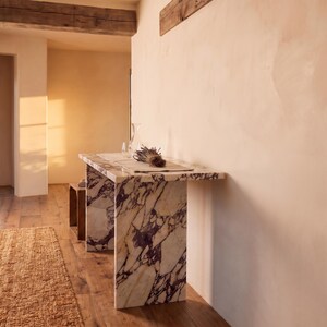 May include: A rectangular console table constructed from white marble with prominent dark purple veining. The table features a flat top surface supported by two vertical slab legs, positioned against a plain wall in a room with wooden flooring.