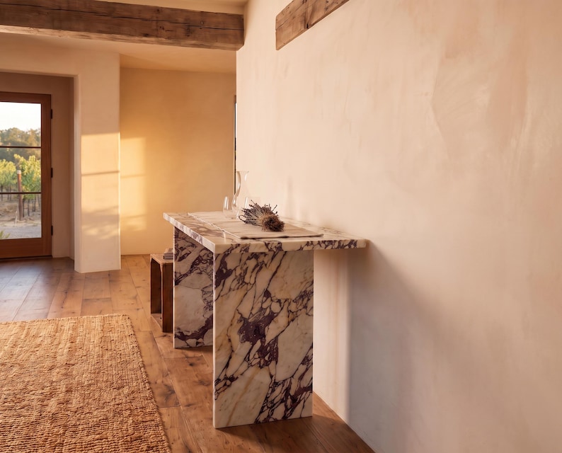 May include: A rectangular console table constructed from white marble with prominent dark purple veining. The table features a flat top surface supported by two vertical slab legs, positioned against a plain wall in a room with wooden flooring.