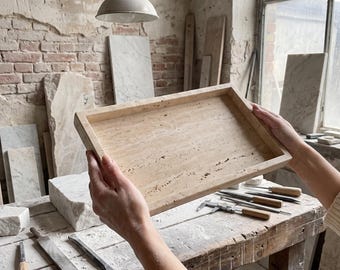 May include: A rectangular tray crafted from beige travertine stone with a natural porous texture and raised edges. The tray features a light tan color with subtle variations in tone and small surface indentations characteristic of the natural stone material.
