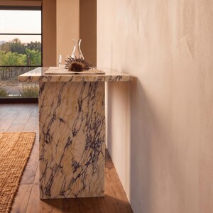 May include: A rectangular console table crafted from cream-colored marble with prominent dark purple veining. The table features a thick, flat top supported by a single solid slab base, displaying a natural stone pattern throughout its entire structure.