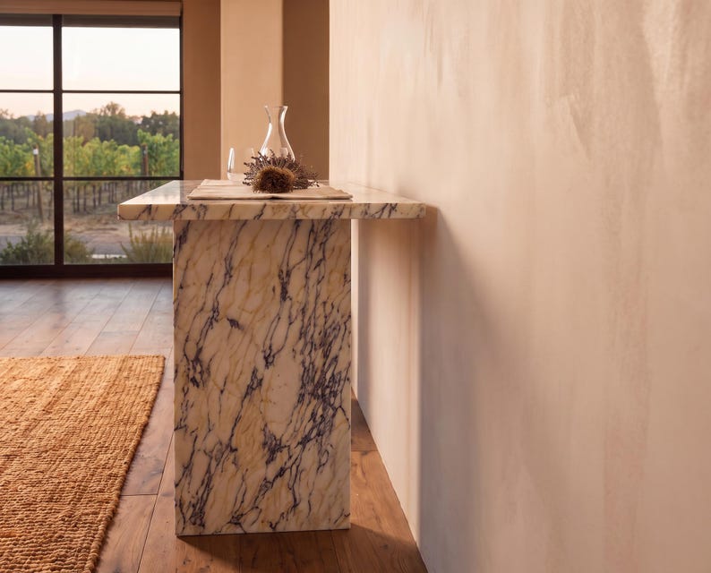 May include: A rectangular console table crafted from cream-colored marble with prominent dark purple veining. The table features a thick, flat top supported by a single solid slab base, displaying a natural stone pattern throughout its entire structure.