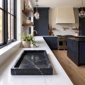 May include: A rectangular black marble tray with white veining sits on a white marble countertop in a kitchen. The kitchen features dark blue cabinetry, a large cream-colored range hood, a stainless steel stove, and a potted green plant.