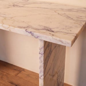 May include: A rectangular console table constructed from white marble with prominent grey and purple veining. The piece features a thick, flat top surface supported by a single vertical slab leg, showcasing the natural stone pattern throughout.