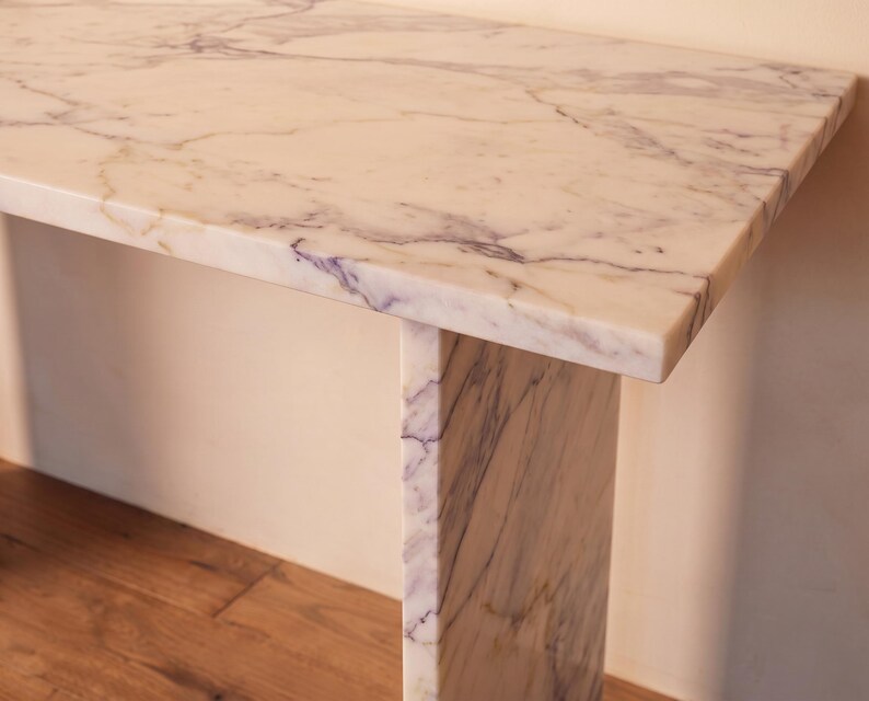 May include: A rectangular console table constructed from white marble with prominent grey and purple veining. The piece features a thick, flat top surface supported by a single vertical slab leg, showcasing the natural stone pattern throughout.