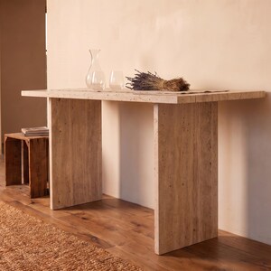 May include: A rectangular console table constructed from beige travertine stone with a porous, textured surface. The table features a thick, flat top supported by two wide, vertical slab legs. A clear glass carafe and glass sit on the table.