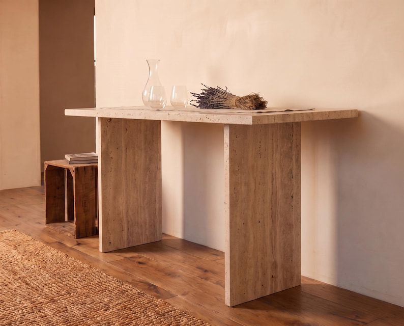 May include: A rectangular console table constructed from beige travertine stone with a porous, textured surface. The table features a thick, flat top supported by two wide, vertical slab legs. A clear glass carafe and glass sit on the table.