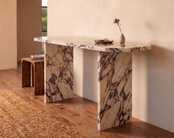 May include: A console table constructed from white marble with prominent purple veining. The table features a long, rectangular top supported by two thick, slab-style legs. A small brass tray and a single dried flower in a vase sit on the surface.