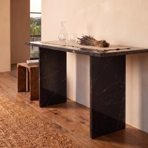May include: A rectangular console table crafted from black marble with white veining, featuring a flat top supported by two vertical slab legs. The table surface holds a clear glass carafe, a glass tumbler, and a bundle of dried lavender on a runner.