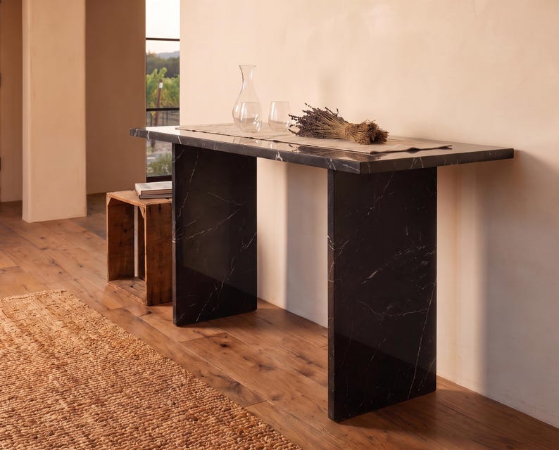 May include: A rectangular console table crafted from black marble with white veining, featuring a flat top supported by two vertical slab legs. The table surface holds a clear glass carafe, a glass tumbler, and a bundle of dried lavender on a runner.