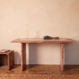 May include: A rectangular wooden console table with a reddish-brown grain pattern, featuring two vertical slab legs. On the table sits a clear glass carafe, two clear glass tumblers, a light-colored runner, and a bundle of dried purple lavender.