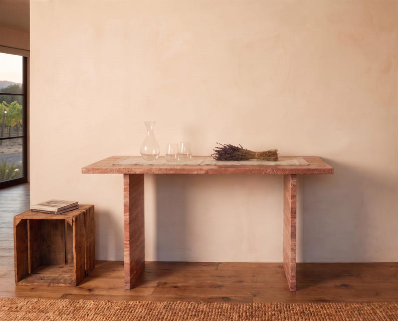 May include: A rectangular wooden console table with a reddish-brown grain pattern, featuring two vertical slab legs. On the table sits a clear glass carafe, two clear glass tumblers, a light-colored runner, and a bundle of dried purple lavender.