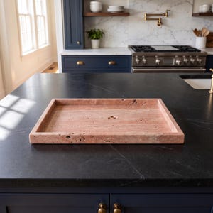 May include: A rectangular tray crafted from pink travertine stone, featuring a natural porous texture and a raised edge. The surface displays the characteristic veining and small indentations inherent to the natural stone material.