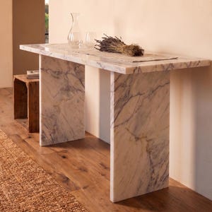 May include: A rectangular console table constructed from white marble with grey veining, featuring a flat top and two vertical slab legs. A clear glass carafe, a glass tumbler, and a dried bundle of lavender are placed on the table surface.