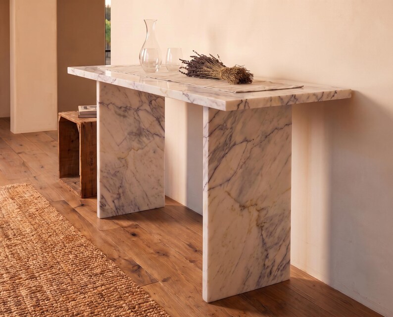 May include: A rectangular console table constructed from white marble with grey veining, featuring a flat top and two vertical slab legs. A clear glass carafe, a glass tumbler, and a dried bundle of lavender are placed on the table surface.