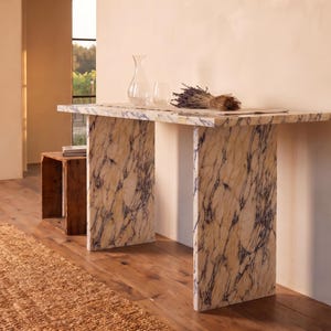 May include: A rectangular console table featuring a cream-colored stone surface with prominent dark purple veining. The table is supported by two matching slab-style legs. On the tabletop sits a clear glass carafe, a glass, and a bundle of dried lavender.