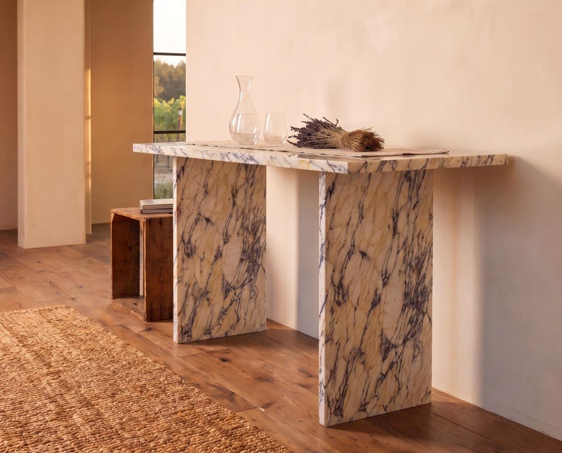 May include: A rectangular console table featuring a cream-colored stone surface with prominent dark purple veining. The table is supported by two matching slab-style legs. On the tabletop sits a clear glass carafe, a glass, and a bundle of dried lavender.