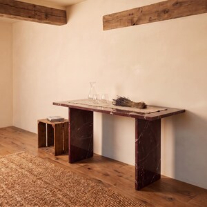 May include: A rectangular console table made of dark red marble with white veining, featuring two slab-style legs. A light-colored runner sits on top, holding a clear glass carafe, two matching glasses, and a bundle of dried lavender stems.