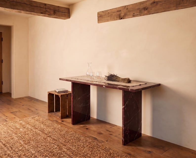 May include: A rectangular console table made of dark red marble with white veining, featuring two slab-style legs. A light-colored runner sits on top, holding a clear glass carafe, two matching glasses, and a bundle of dried lavender stems.
