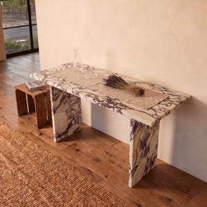 May include: A rectangular console table crafted from white marble with prominent purple veining. The table features a flat top and two slab-style legs. A beige fabric runner, a clear glass carafe, and a bundle of dried lavender rest on the surface.