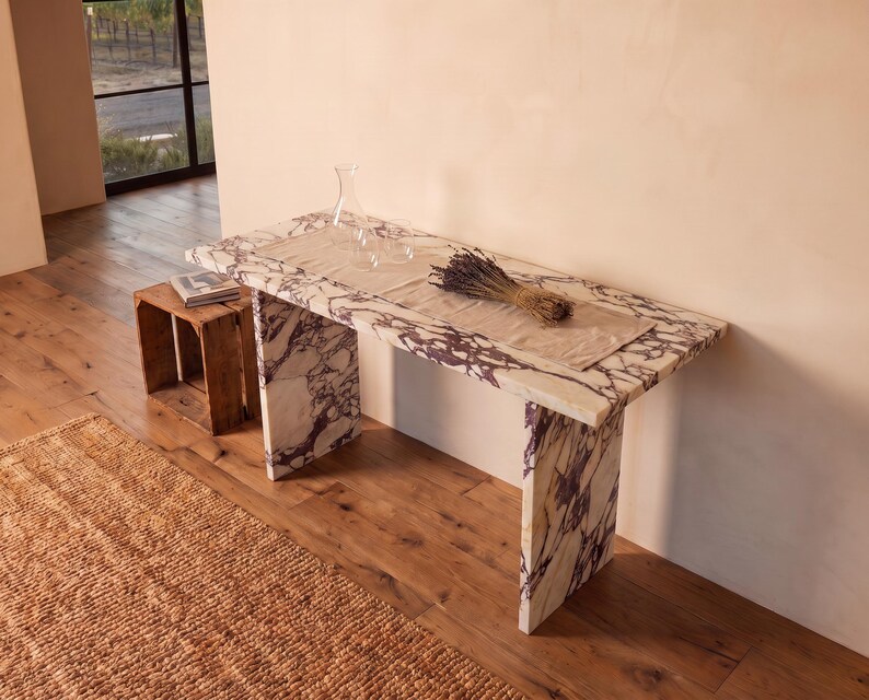 May include: A rectangular console table crafted from white marble with prominent purple veining. The table features a flat top and two slab-style legs. A beige fabric runner, a clear glass carafe, and a bundle of dried lavender rest on the surface.