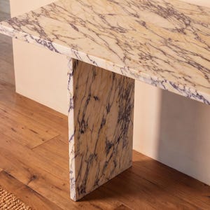 May include: A console table constructed from cream-colored marble with prominent, dark purple veining throughout the surface and the single vertical support leg. The piece features a rectangular top and a flat, slab-style base design.
