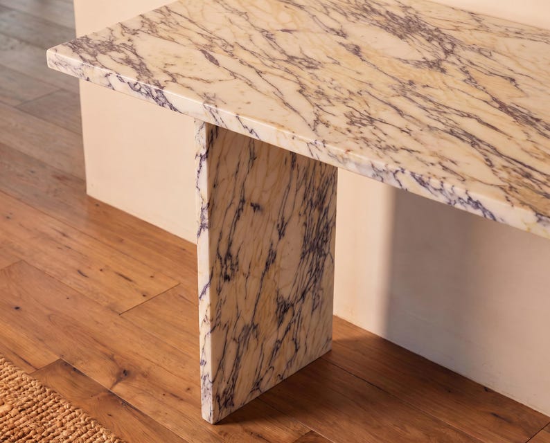 May include: A console table constructed from cream-colored marble with prominent, dark purple veining throughout the surface and the single vertical support leg. The piece features a rectangular top and a flat, slab-style base design.