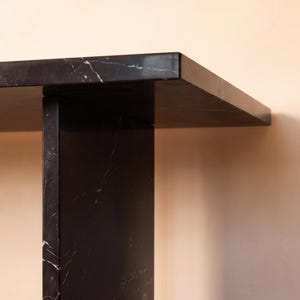 May include: A black marble console table featuring a smooth, polished surface with subtle white veining. The structure consists of a thick, rectangular tabletop supported by a vertical, flat slab base, showcasing a clean, geometric design.