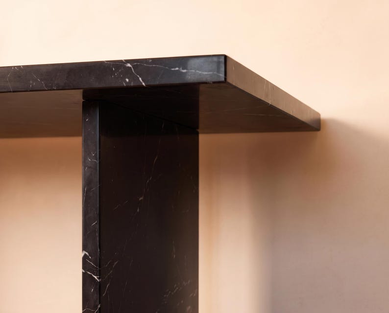May include: A black marble console table featuring a smooth, polished surface with subtle white veining. The structure consists of a thick, rectangular tabletop supported by a vertical, flat slab base, showcasing a clean, geometric design.