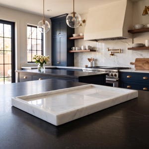 May include: A rectangular white marble tray with a smooth, polished surface and raised edges. The tray features subtle grey veining throughout the stone, showcasing a clean, flat design suitable for serving or display on a dark kitchen countertop.
