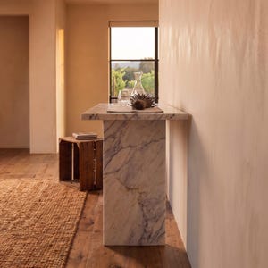 May include: A rectangular white marble console table with grey veining, featuring a thick top and a solid vertical base. The table surface holds a clear glass carafe and a dark brown dried flower head, positioned against a light-colored wall.