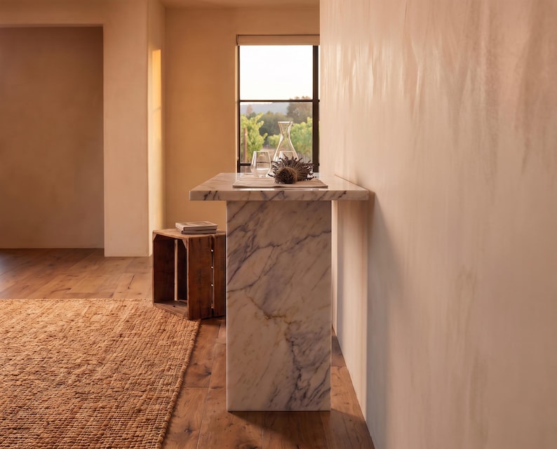 May include: A rectangular white marble console table with grey veining, featuring a thick top and a solid vertical base. The table surface holds a clear glass carafe and a dark brown dried flower head, positioned against a light-colored wall.