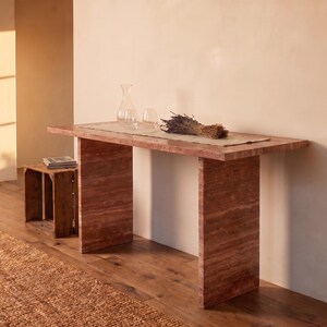 May include: A rectangular console table made of reddish-brown travertine stone with a smooth top and two vertical slab legs. On the table sits a clear glass carafe, a clear glass tumbler, and a bundle of dried lavender stems on a runner.