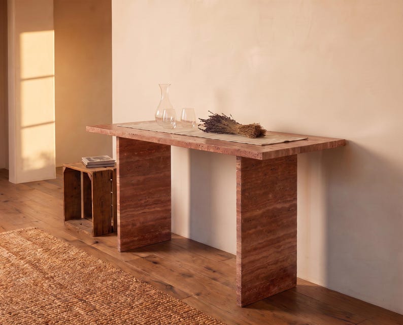 May include: A rectangular console table made of reddish-brown travertine stone with a smooth top and two vertical slab legs. On the table sits a clear glass carafe, a clear glass tumbler, and a bundle of dried lavender stems on a runner.