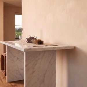May include: A white marble console table with gray veining, featuring a rectangular top and two slab legs. On the table sits a light-colored fabric runner, a clear glass carafe, a clear glass tumbler, and a dried bundle of purple lavender flowers.