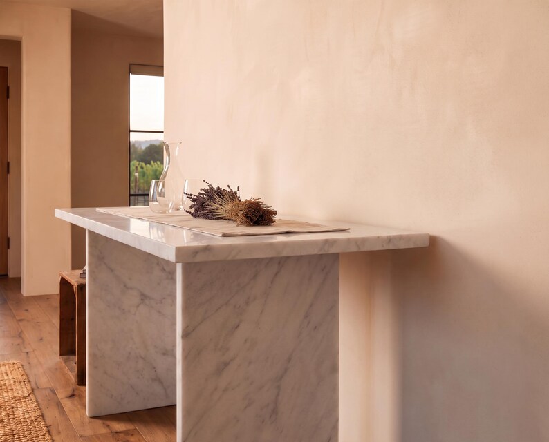 May include: A white marble console table with gray veining, featuring a rectangular top and two slab legs. On the table sits a light-colored fabric runner, a clear glass carafe, a clear glass tumbler, and a dried bundle of purple lavender flowers.
