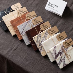 May include: Eight rectangular stone tiles arranged in a row, each with a brown paper tag labeled 01 through 08. The stones include Toros Black, Beige Travertine, Red Travertine, Lilac Marble, Rosso Marble, Violet Marble, White Marble, and Calacatta Viola.