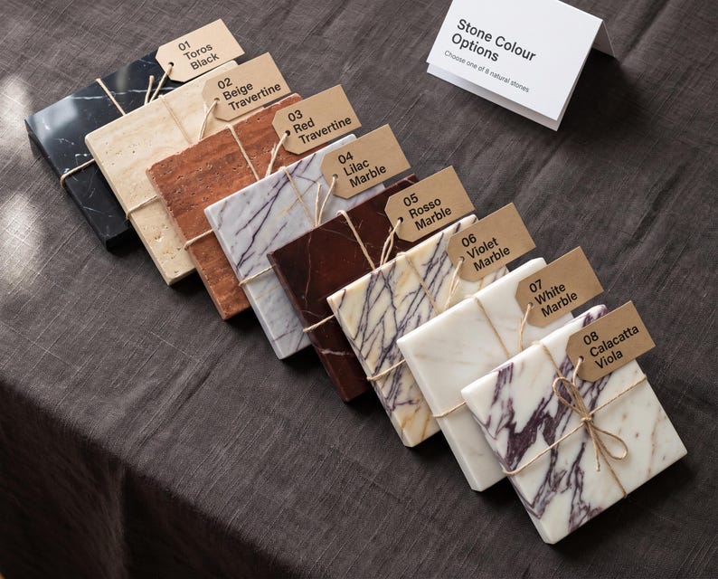 May include: Eight rectangular stone tiles arranged in a row, each with a brown paper tag labeled 01 through 08. The stones include Toros Black, Beige Travertine, Red Travertine, Lilac Marble, Rosso Marble, Violet Marble, White Marble, and Calacatta Viola.