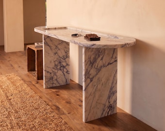 May include: A white marble console table with prominent purple veining, featuring an elongated oval top with a recessed surface and two vertical slab legs. A small set of metal keys and a folded dark brown fabric item rest on the tabletop.