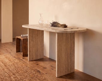 May include: A beige travertine console table with a smooth oval top and two vertical slab legs. On the table sits a clear glass carafe, a glass tumbler, and a bundle of dried lavender resting on a rectangular fabric runner.